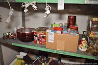 Items on green metal shelving including large red glass bowl, boxes of Christmas wrapping paper, tins, ribbons, brass bells, and a snowman decoration