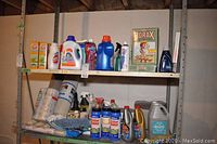 Wider view of two metal shelves filled with assorted cleaning products: laundry detergents, sprays, boxed baking soda, Borax, shelf liner roll, dusters, drain cleaner.