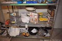 Photo showing upper and lower shelf with boxed Joe Max cookie press, white plastic dish rack, clear plastic tiered cake stands, yellow lidded small dishes, and various metal bakeware including springform pans and pie pan.