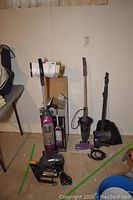 Photo showing four vacuums: two upright Shark vacuums, a Hoover stick vacuum, and a black canister vacuum placed on a concrete floor against a white wall.