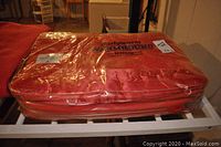 Earlywarm Warmbound red wool blanket folded inside original zipped plastic packaging sit on a white rack.