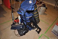 Blue backpack child carrier with attached frame and harness system, red and black hand pump beside it, black and yellow carry bag, and navy blue folding chair seen from the side with orange mini basketball in front.