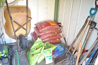 Photo shows rusty metal wheelbarrow, several bags of garden mulch and soil, and a variety of long-handled garden tools including a snow shovel, flat shovel, garden fork, and pick tool.