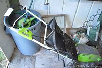 Overview of the Greenworks electric lawn mower with black grass catcher bag placed next to a large gray plastic trash can holding garden accessories.