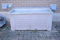 Front view of beige plastic deck storage box showing horizontal panel design, lid closed, situated outside on brick patio against brick wall; visible discoloration and dirt on surface.