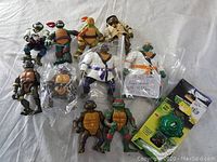 All figures laid flat on a white surface showing various Teenage Mutant Ninja Turtles action figures including some in plastic bags and a projectile toy in packaging.