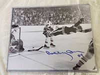front view of 8x10 black and white photo featuring Bobby Orr mid-air over ice near goal, signed in blue ink lower center 'Bobby Orr #4'.