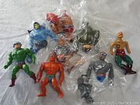 Photo showing 8 Masters of the Universe action figures, some in plastic bags, arranged on a flat surface