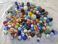 Wide view of a large assortment of glass marbles in various colors and swirl designs laid out on a white cloth.