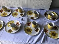 Eight dessert plates arranged with matching coffee mugs on a white tablecloth, showing floral patterns and mug colors.