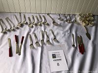 Full lot laid out on white cloth showing silver spoons, forks, knives, red handled butter knives, serving utensils, and multiple small napkin holders.