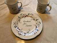 Plate and two mugs with Christmas themed text and holly decoration shown on beige surface.