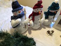 Three snowman plush figures wearing knitted hats and scarves, each holding wooden sticks.