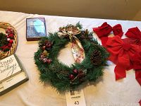 Photo showing a green Christmas wreath decorated with pine cones, berries, and a floral bow, several large red Christmas bows, a smaller wreath with a sign, and a box of Christmas cards.
