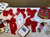 Wide shot of 7 large red velvet Christmas bows, assorted ribbon spools, folded Christmas-themed vinyl tablecloth with snowman pattern, and boxed Christmas cards on beige surface.
