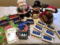 Full assortment of holiday plush gift bags and GE and Westinghouse exterior Christmas light bulbs on table including three plush holiday characters (Santa, snowman, moose) and multiple packs of bulbs.
