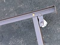 Close-up photo of metal bed frame corner showing bolt and support bracket with dark brown finish and slight rust.