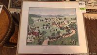 Framed print of a colorful rural village scene with houses, people, animals, mountains, in a gold-tone frame with cream matting.