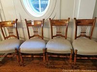 Four wooden framed chairs with cushioned beige seats, arranged side-by-side against a wall below a round window, showing wear on the cushions.