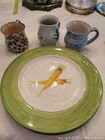 Three ceramic decorative mugs and one hand painted plate with illustration of carrots and green rim, displayed on a table.