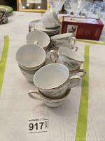 Eleven white Mikasa tea cups stacked and arranged on a table showing decorative gold rims and gray floral patterns, some wear visible on the gold trim.