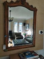 Front view of the large hand carved frame mirror with beveled glass hanging on the wall in a living space showing reflective surface capturing furniture and windows.
