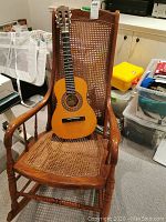 Photo showing wooden rocking chair with cane seat and backrest, child's Montanna guitar resting on it, surrounded by household items.