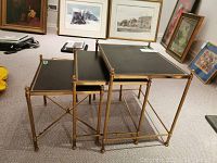 Three brass and leather nesting tables aligned from smallest to largest on carpeted floor with framed artwork in background