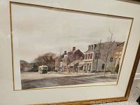 Framed print showing street scene with buildings, bus, and winter trees. Frame shows visible wear and print has shifted inside frame.