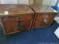 Front and slightly angled view of two fruitwood bedside tables on blue carpet showing drawer and cabinet doors, surface wear visible