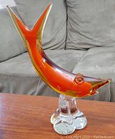Side view of orange and yellow Murano glass fish sculpture on clear glass base on wooden surface with couch in background.