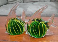 Photo of two green and yellow striped glass fish figurines on a wooden surface, showing clear glass fins and roughly 4 inches tall.