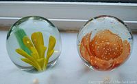 Two clear glass spherical paperweights side by side on a windowsill. The left paperweight contains yellow flowers with green stems. The right paperweight has an orange rose-like flower with bubbles around it.