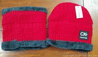 Photo of the red knit hat and matching neck warmer laid flat on a wooden surface. Both pieces have gray faux fur lining. The hat has a Calvin Klein logo patch.