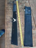 Photo of Eagle Claw machete laying flat with measuring tape showing approximately 18 inch blade length and black nylon sheath beside it.