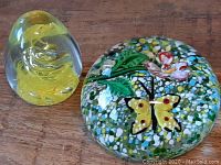 Two glass paperweights shown together on wooden surface, one egg-shaped with a yellow spiral and the other round with butterfly floral print.