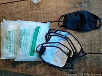 Photo of two packs of translucent disposable plastic food prep gloves partially visible with some packaging text. Five white cloth masks with black trim and one black cloth mask laid out showing variety in the set.