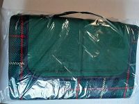 Folded picnic blanket showing green tartan plaid pattern with red and beige lines wrapped in plastic, highlighting the cloth side and the carry handle.