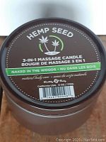 Top view of brown metal container lid with Hemp Seed label showing 3-in-1 Massage Candle, scent "Naked in the Woods", 6 oz/170g.