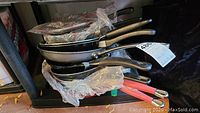 Stacked skillets and pans with visible black handles and protective plastic wrapping hard to count individual items clearly.