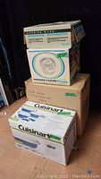 Stacked boxes showing Cuisinart ceramic stoneware set box at bottom with Corning Ware divided dish and Mouli spin-dry and Wolfgang Puck tri-grill boxes stacked above
