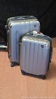 Two metallic gray hard shell suitcases standing upright with chrome corner protectors and spinner wheels.
