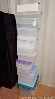 Stack of multiple clear plastic storage containers of various sizes and styles, predominantly rectangular with some having blue or purple lids, others lidless.