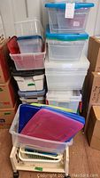 Stacked various plastic storage bins and containers with lids in different colors and sizes, a clear basket holds smaller red and clear bins.