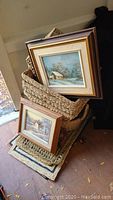 Three wicker baskets stacked with two framed winter landscape paintings leaning in front and on top of them, also showing some woven floor mats beneath.