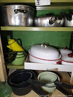 Two shelves with multiple pots and pans, including large aluminum pots on the top shelf, green enamel saucepans with black handles, and a yellow enamel coffee pot on the lower shelf.
