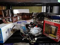 Overview of shelf with various fry pans, roasters, Pyrex stove tops, asparagus steamer and Meyer roaster boxed and unpacked.