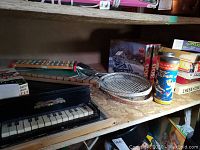 View of one shelf showing vintage toys and games including a black toy piano, two xylophones, wooden tennis racquets, and boxed board games