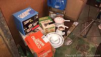 Photo showing boxed Corning Ware cookware and Corelle Cornflower dishes including cups, saucers, plates, and a covered casserole pan set.