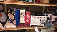 Shelf showing boxes of kitchen appliances and accessories including KitchenAid stand mixer bowl and combi whip, food chopper, Wiltshire knife block, and various kitchen tools.